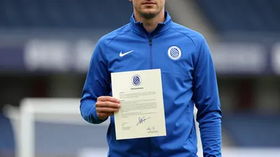 A mid-30s anonymous Cercle Brugge staff member in a tight waist-up shot, wearing a solid blue training jacket, holding a...