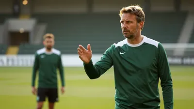 A 35-year-old Cercle Brugge coach with a weathered complexion, wearing a plain dark green and white crew-necked polyester...