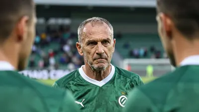 A close-up shot of a middle-aged, ruggedly-built, unshaven head coach in his late 50s, with a stern expression, gazing...