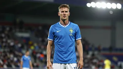 A 29-year-old Swedish striker with a determined expression, wearing a blue jersey with a white collar, and a white pair...