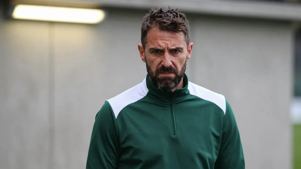 A medium shot of a rugged, 35-year-old, bearded assistant coach, clad in a dark green and white polyester training top...