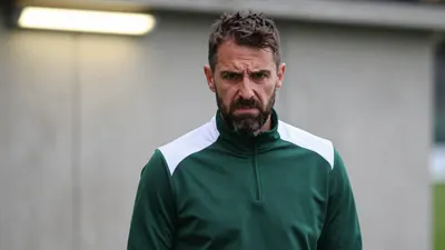 A medium shot of a rugged, 35-year-old, bearded assistant coach, clad in a dark green and white polyester training top...