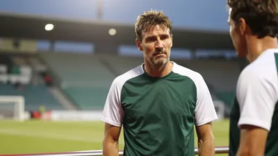 A medium shot of a rugged, 35-year-old coach, wearing a dark green and white plain cotton training top, his face set in a...