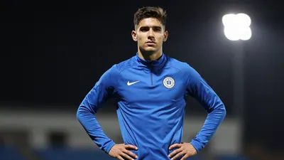 A 20-year-old midfielder in a solid blue training kit, standing confidently with hands on hips, 85mm f/1.4 shallow depth...