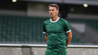 A 35-year-old, ruggedly-built assistant coach in dark green and white plain training top, with white collar and cuffs...