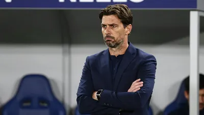 A 35-year-old male football manager in a dark blue suit, standing in a dugout with a serious expression, arms crossed...