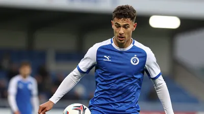 A photorealistic image of a 22-year-old Cercle Brugge midfielder in a blue jersey with white sleeves, playing with a ball...
